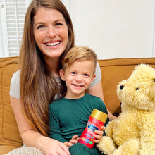 Woman and child sitting on a couch with a teddy bear and a container ofvitamins.